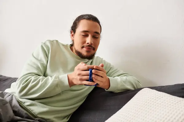 A young man sits comfortably on a couch, sipping from a warm mug, enjoying a peaceful moment. — Stock Photo