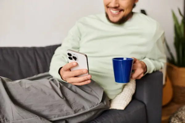 Ein junger, gutaussehender Mann lümmelt auf einer Couch und nippt an einem Drink, während er sein Handy zu Hause benutzt. — Stockfoto