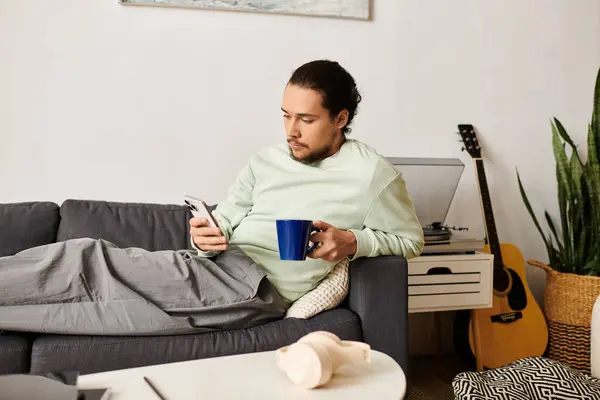 Comfortably lounging on the couch, a young man enjoys a drink while browsing on his phone. — Stock Photo