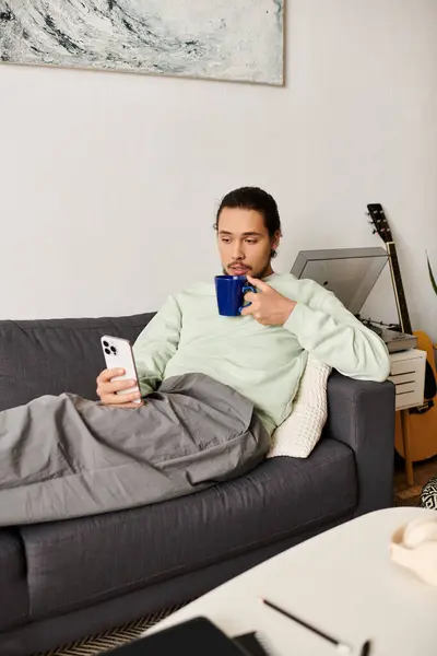 A handsome young man relaxes on a sofa, sipping a drink and browsing his smartphone. — Stock Photo