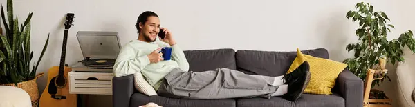 Un joven guapo descansa en un sofá, charlando por teléfono y bebiendo café cómodamente. - foto de stock