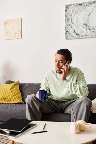 Un giovane uomo si rilassa a casa, sorseggiando un caffè e chiacchierando al telefono. — Foto stock