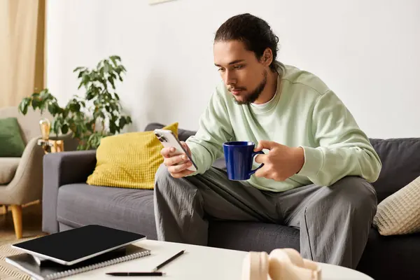 Un bel giovanotto si rilassa sul divano, legge sul telefono e sorseggia un drink caldo. — Foto stock