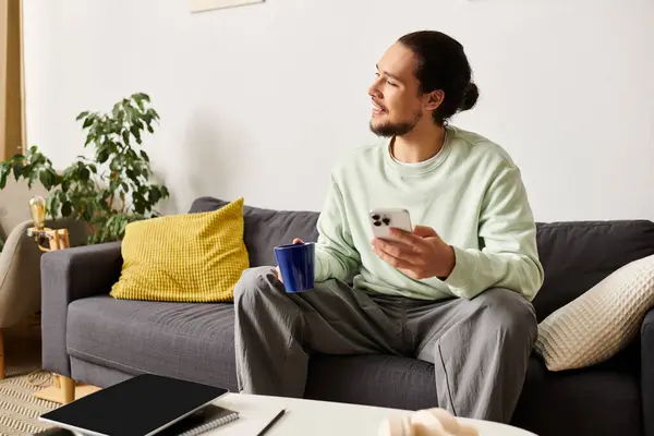 El hombre se relaja en casa, bebiendo una bebida caliente y usando su teléfono inteligente. - foto de stock