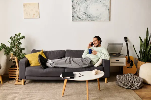 Un bel giovane uomo si rilassa su un divano a casa, chiacchierando al telefono e sorseggiando un caffè. — Foto stock