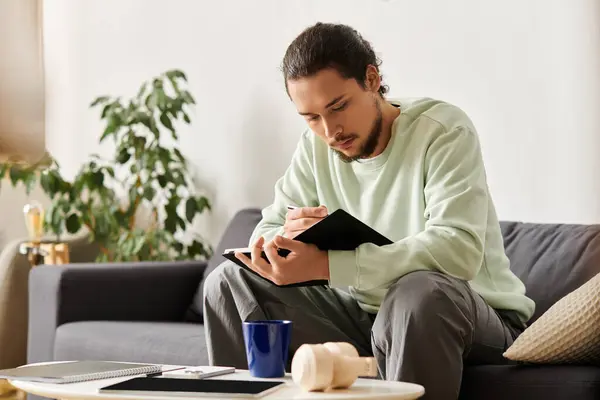 Focused young man writes in a notebook while seated comfortably at home, enjoying his surroundings. — Stock Photo