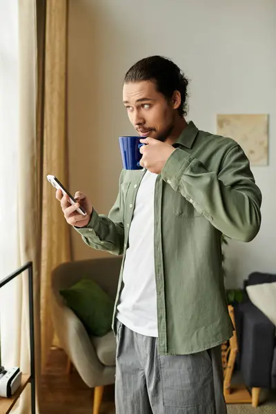 Habitación luminosa con un joven bebiendo té de una taza azul mientras mira su teléfono. — Stock Photo