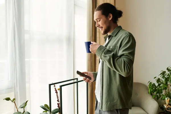 Un hombre joven y guapo está en una habitación acogedora, bebiendo de una taza azul mientras usa su teléfono. — Stock Photo