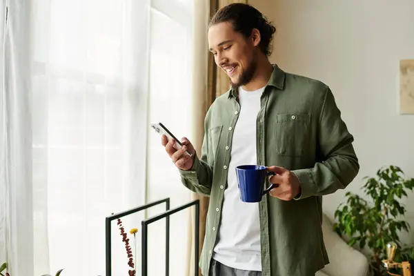 Un giovane uomo sta sorridendo mentre controlla il suo telefono, tenendo una tazza blu nel suo accogliente spazio domestico. — Foto stock