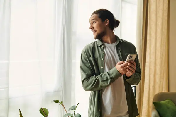 A young and attractive man stands near a window, casually gazing out while holding his phone. — Stock Photo