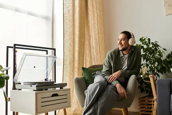 Joven guapo se sienta cómodamente en una silla acogedora, escuchando música con auriculares. - foto de stock
