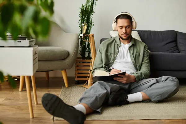 A young man sits comfortably on the floor, writing in his notebook while wearing headphones. — Stock Photo