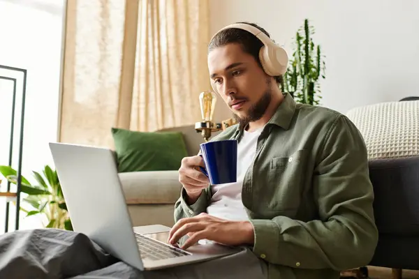 A young man sits at home, sipping coffee and working intently on his laptop. — Stock Photo
