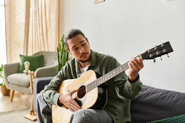 Ein junger, gutaussehender Mann spielt seine Gitarre, während er gemütlich in einer entspannten häuslichen Umgebung sitzt. — Stockfoto