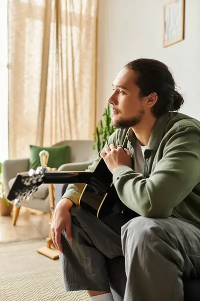 Handsome young man sits comfortably at home, deep in thought while holding his guitar. — Stock Photo