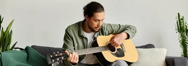 Un jeune homme beau s'assoit confortablement à la maison, jouant habilement de la guitare et appréciant sa musique. — Photo de stock
