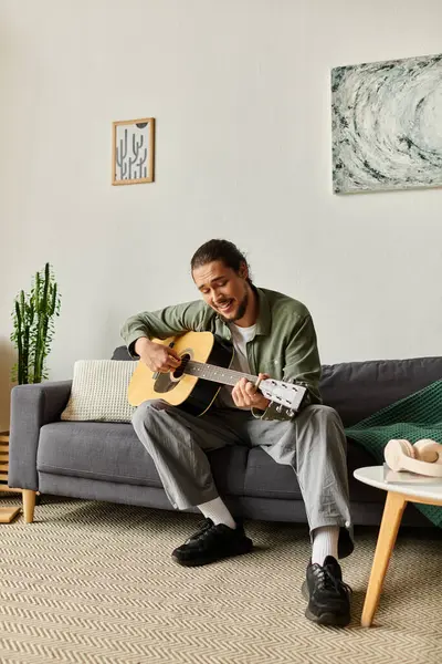 A young man joyfully plays guitar on the sofa, filling his cozy space with cheer. — Stock Photo
