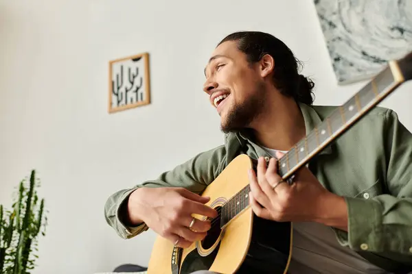 A handsome young man enjoys playing his guitar at home, filled with a relaxed and cheerful mood. — Stock Photo
