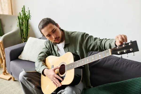 Un hombre joven y guapo sintoniza su guitarra mientras está sentado cómodamente en su elegante casa. - foto de stock