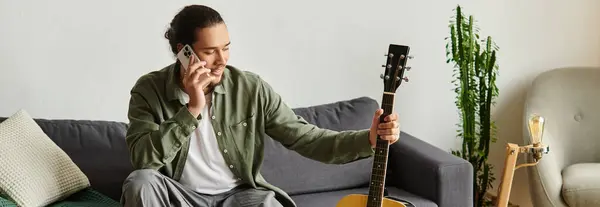 Un joven guapo se relaja en casa, charlando alegremente por teléfono con una guitarra. - foto de stock