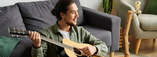 A young man sits comfortably in his living room strumming a guitar, immersed in music. — Stock Photo