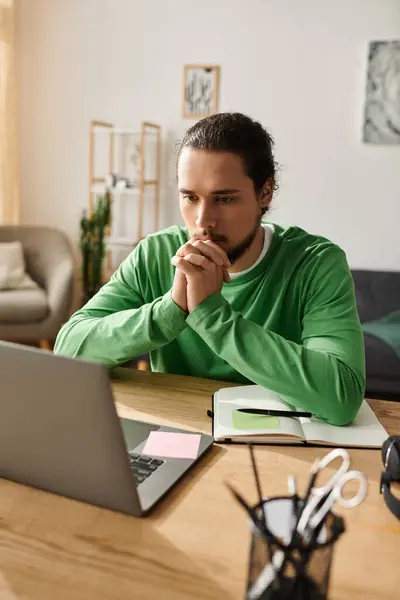 Concentrated young man in a green shirt sits at a wooden desk working on his laptop at home. — Stock Photo