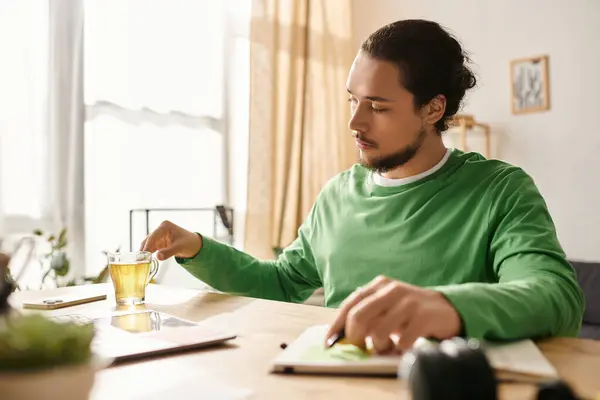 Un giovane uomo in camicia verde sorseggia tè, concentrato sul suo lavoro a casa durante la luce del sole pomeridiana. — Foto stock