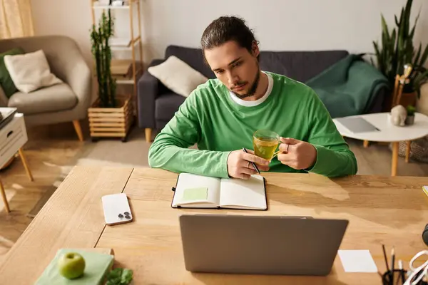 Un joven bebe té y escribe en un cuaderno en una acogedora mesa de madera. - foto de stock