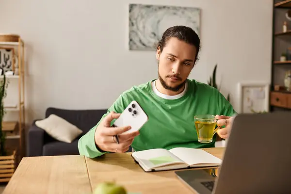 Handsome young man relaxed at home, sipping tea and engaging with his smartphone, focused and calm. — Stock Photo