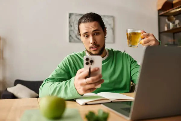 A young handsome man sits at a table with a laptop, sipping tea and checking his phone. — Stock Photo