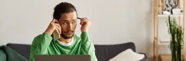 A young man with a thoughtful expression uses his phone while seated at his desk in a cozy home. — Stock Photo