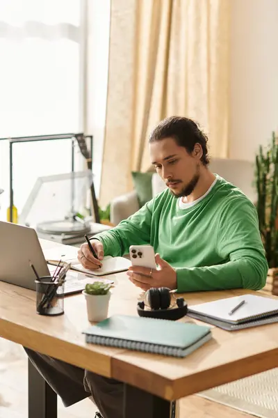 A young man sits at a modern desk in his home, focused on his smartphone while writing notes. — Stock Photo