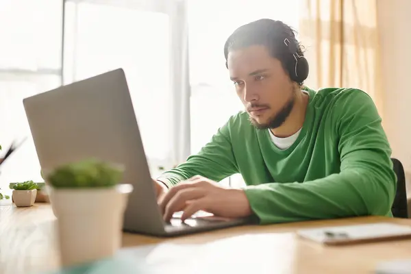 Determined young man works intently on his laptop at home, wearing headphones amidst natural light. — Stock Photo