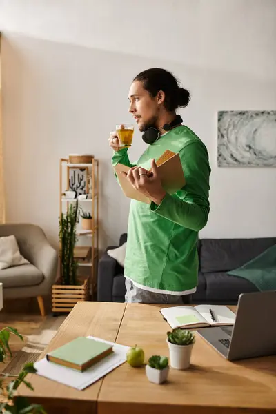 Un giovane uomo in camicia verde sta a casa sorseggiando tè e tenendo libri in un ambiente rilassato. — Foto stock