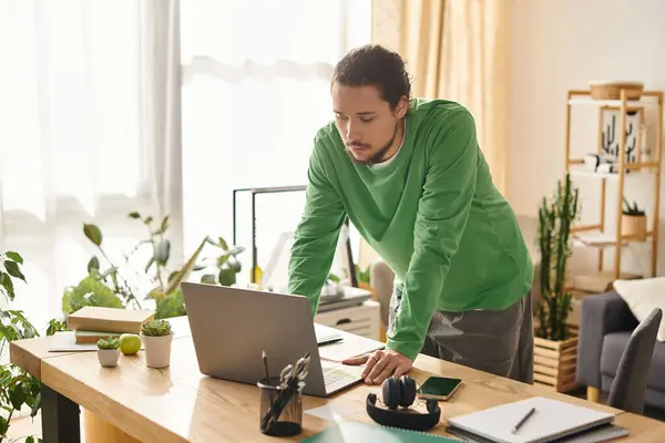A young man works on his laptop at a wooden table in a cozy, green filled home. — Stock Photo