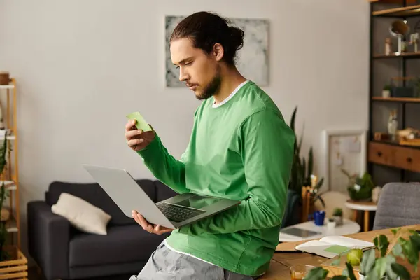 Focused young man in a green shirt reviews notes while using a laptop in his stylish home setting. — Stock Photo