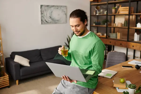 Relaxed young man in a green sweater smiles at his laptop while sipping tea in a sunny room. — Stock Photo