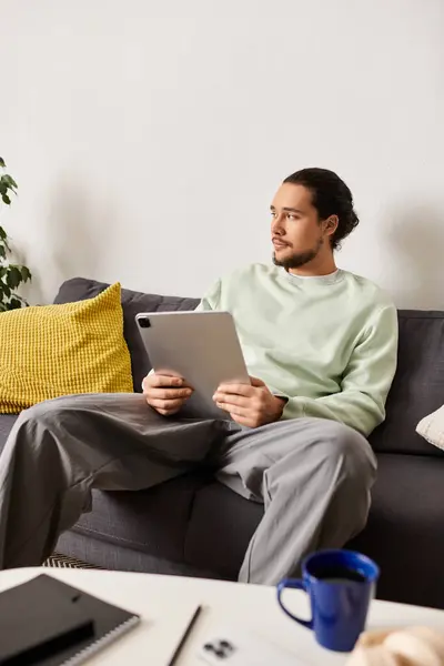 A young man sits comfortably on a couch, engaged with his tablet in a warm living space. — Stock Photo