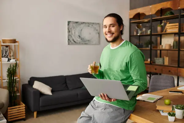 A cheerful young man in a green shirt holds a cup of tea while using his laptop at home. — Stock Photo