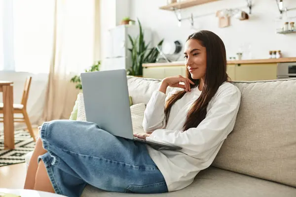 Beautiful woman relaxes on the couch, engaged with her laptop in a cozy home environment. — стоковое фото
