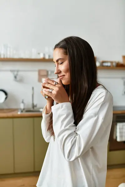 Woman with long hair smiles gently while holding a warm drink in her charming kitchen. — Stock Photo