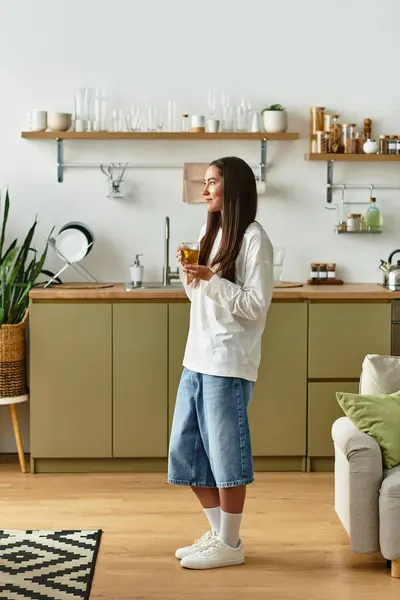 A young beautiful woman stands in her cozy kitchen, savoring a drink in a relaxed moment. — Stock Photo