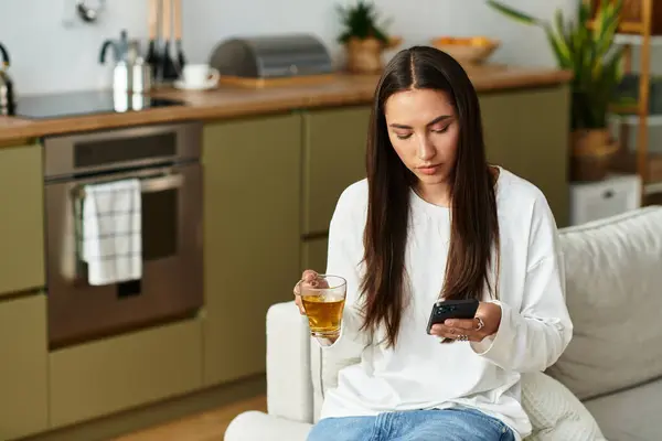 Pretty young woman relaxes at home, sipping tea while engrossed in her mobile phone. — Stock Photo