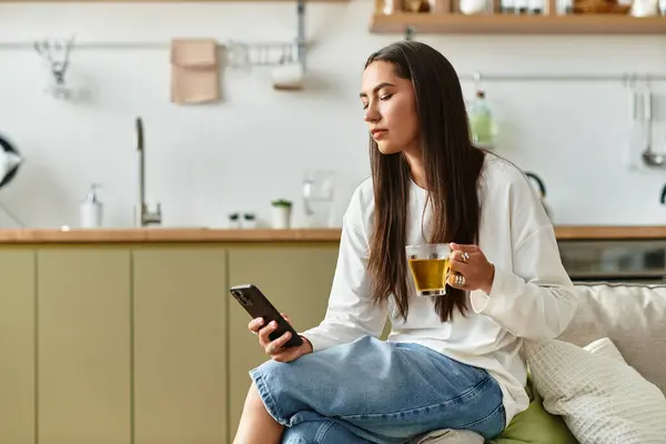 A young woman relaxes at home, sipping tea while engrossed in her smartphone, feeling cozy. — Stock Photo