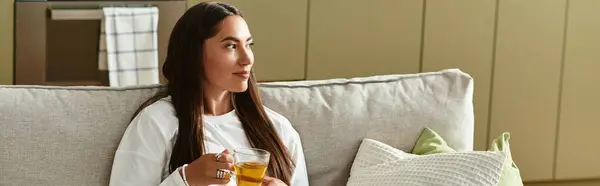 A young beautiful woman sits comfortably at home, savoring a warm drink while relaxing. — Stock Photo
