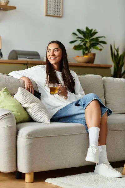 Young woman sits on a cozy sofa, sipping a refreshing drink while relaxing at home. — Stock Photo