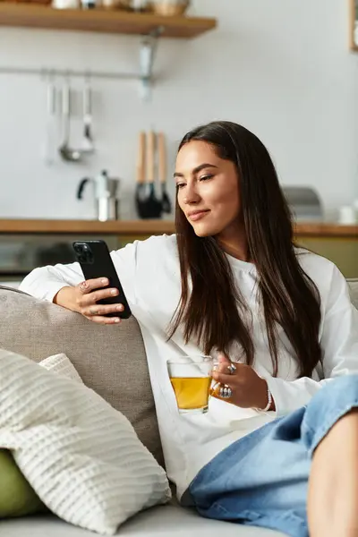 A young woman relaxes on a couch, sipping tea and browsing her phone at home. — Stock Photo