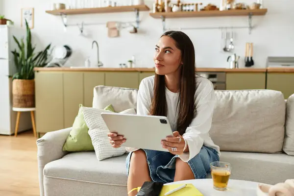 Pretty woman relaxes comfortably in her cozy living room, engaged with her tablet while sipping tea. — Stock Photo