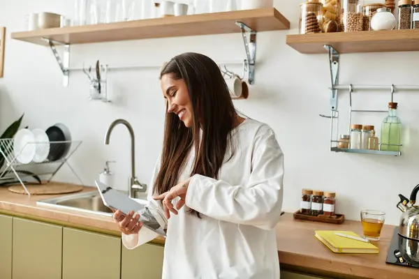 A cheerful young woman smiles as she uses her tablet in a cozy kitchen, surrounded by warm decor. — Stock Photo