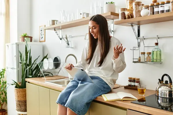 A beautiful young woman relaxes in her kitchen, happily interacting with her tablet and sipping tea. — Stock Photo